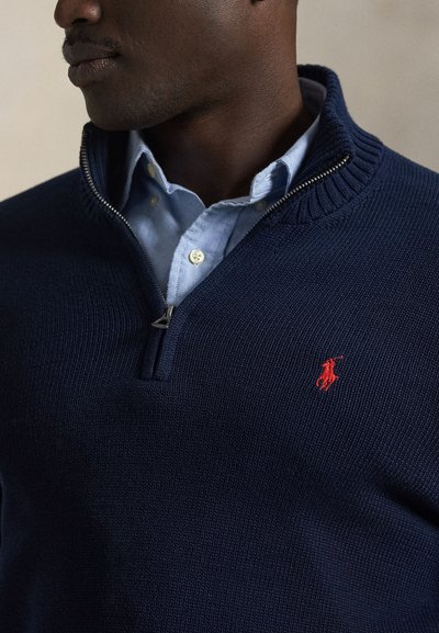 Tamsiai mėlynas megztinis su užtrauktuku ir ribbiniu apykakle, su raudonu logotipu ant krūtinės. Dėvimas ant šviesiai mėlynos marškinių su apykakle, demonstruojantis sluoksniuotas tekstūras.