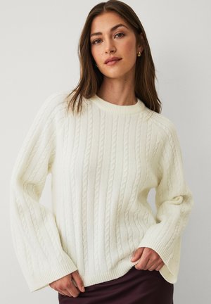 Young woman with long brown hair wears a white cable knit sweater and dark skirt, posing against a plain light background.
