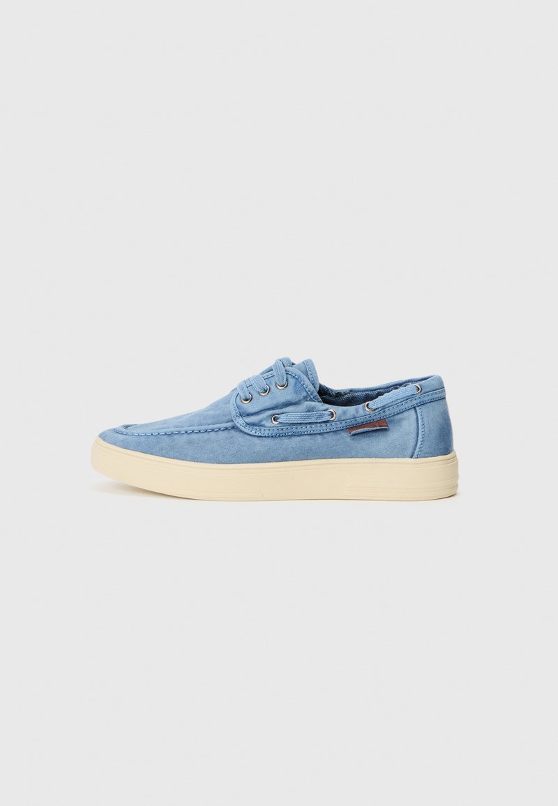 Zapatilla de lona azul claro con un diseño de bajo perfil, punta redonda y suela de goma crema. Presenta ojales y un detalle de costura en la parte superior.