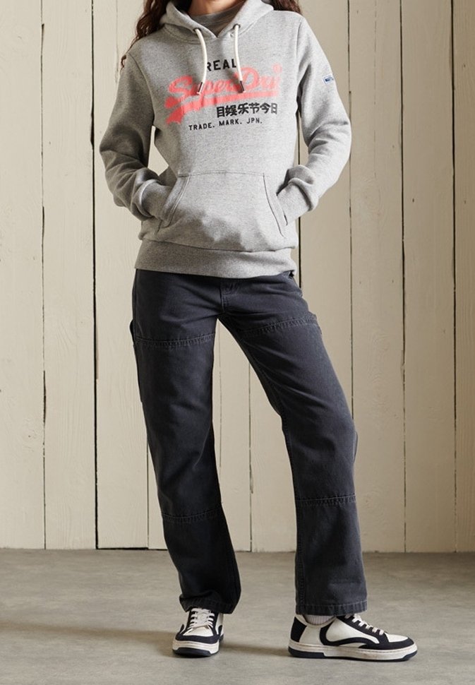 Personne portant un sweat à capuche gris Superdry, un pantalon large foncé et des baskets noires et blanches, debout contre un mur en bois clair.
