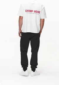 Hvid t-shirt med korte ærmer, der har teksten "CHAMP MODE" i rød på ryggen, parret med sorte joggers og hvide sneakers.