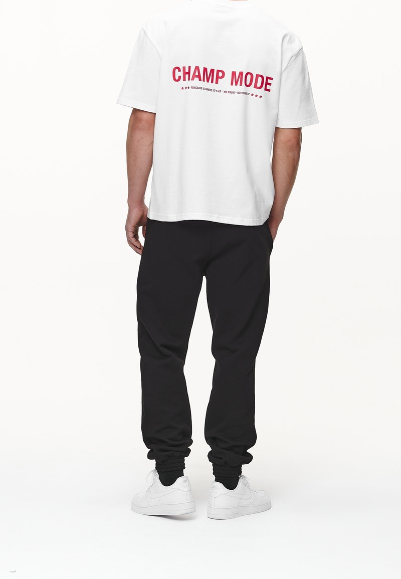 Hvid t-shirt med korte ærmer, der har teksten "CHAMP MODE" i rød på ryggen, parret med sorte joggers og hvide sneakers.