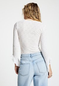 Weißes Langarmshirt mit strukturierter Stoffqualität und Rüschensaum an den Ärmeln, kombiniert mit hellblauen, hoch taillierten Jeans.