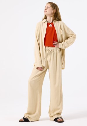 Pantalones largos de lino beige y una camisa de gran tamaño a juego se combinan con un top corto de canalé rojo. Zapatos negros sin cordones cuentan con acentos decorativos.