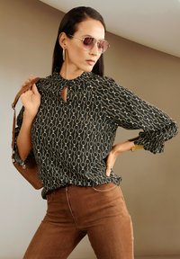 Schwarze Bluse mit goldenen Kreismustern, Ausschnitt mit Schlüsselöffnung und Rüschendetails, kombiniert mit braunen eng anliegenden Jeans und einer braunen Handtasche. Sonnenbrillen vervollständigen den Look.