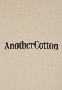 Beige stof med broderet tekst "AnotherCotton" i sort, lavet med en enkel, moderne skrifttype med en let hævet tekstur.