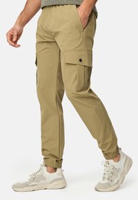 Kaki cargo bukser med elastiske manchetter, der har sidelommer og lukning med knap foran. Båret med teksturerede, lyse sneakers.