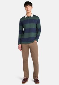 Camisa polo de manga larga a rayas verdes y marino con un cuello beige, combinada con pantalones caqui y zapatos marrones. Ajuste casual, tejido textil.