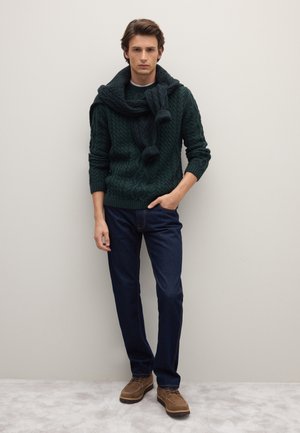 Suéter de punto trenzado verde oscuro con una bufanda a juego caída sobre los hombros, combinado con jeans azul oscuro y zapatos marrones de cordones.