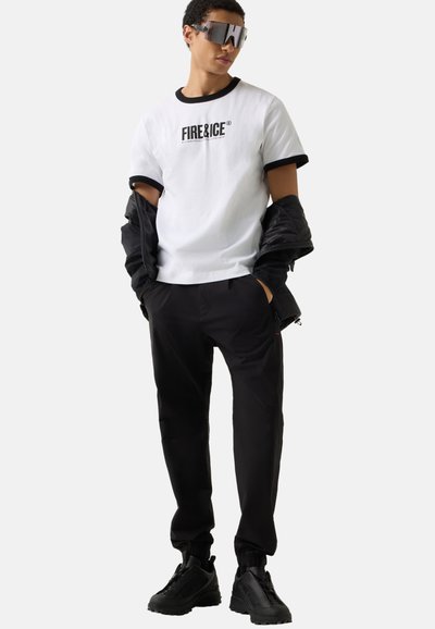 Ung mand iført en hvid Fire&Ice t-shirt, sort jakke hængende over armene, sorte bukser, chunky sorte sneakers og refleksive store solbriller.