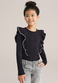 Top lungo a maniche nere a costine con dettagli arricciati bianchi lungo le spalle. Abbinato a pantaloni in denim grigio. Sfondo semplice.