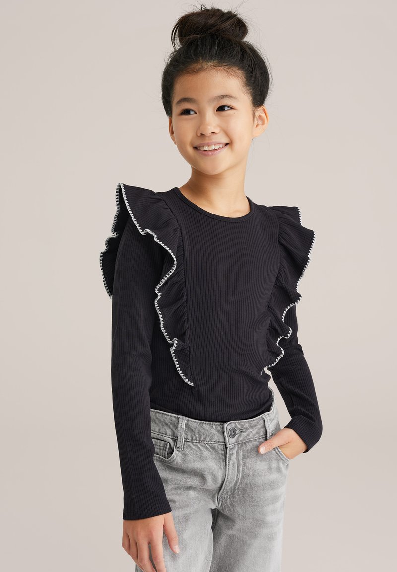 Top lungo a maniche nere a costine con dettagli arricciati bianchi lungo le spalle. Abbinato a pantaloni in denim grigio. Sfondo semplice.