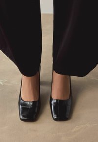 Czarne lakierowane szpilki na kwadratowym nosku z subtelnym wytłaczanym wzorem krokodyla, zestawione z szerokimi czarnymi spodniami na beżowym tle.