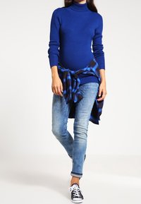 Suéter de cuello alto azul con textura acanalada, combinado con jeans azul claro y una camisa a cuadros negra y azul anudada a la cintura. Zapatillas negras.