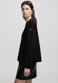 Sweatshirt noir avec des détails tressés sur les deux manches, une coupe décontractée, des manches longues et une texture douce. Associé à une jupe en cuir.