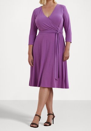 Femme portant une robe portefeuille violette longueur genou avec manches trois-quarts et des sandales noires à talons hauts, debout devant un fond uni.