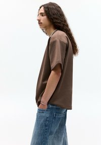 Tricou din bumbac maro cu mâneci scurte, croială lejeră și decolteu rotund, asortat cu pantaloni din denim albastru deschis cu buzunare aplicate.
