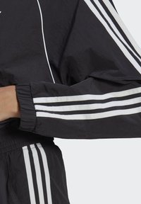 Giacca a vento nera con design a tre strisce bianche sulle maniche e sui polsini, con orlo elasticizzato arricciato e tessuto leggero.