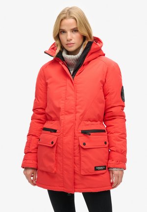 Red padded parka jacket with a hood, two front pockets, a black zipper, and a logo patch on the lower left side. Features a textured gray sweater underneath.