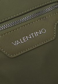 Tessuto in nylon verde oliva con superficie strutturata, dotato di chiusura con zip argentata e un accentuato logo "VALENTINO" argentato.