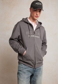 Grå hætte sweatshirt med frontlommer, prydet med teksten "BIG STAR AUTHENTIC". Bæres over en lyserød polo og kombineret med lyseblå jeans.