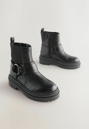 Stivali alla caviglia in pelle nera con zip laterale, cinturino regolabile e suola in gomma texturizzata; presentano dettagli cuciti e un tacco robusto.