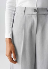 Gray, pleated trousers with a button closure at the waistband, shown with a hand resting on the fabric. The texture appears smooth and tailored.