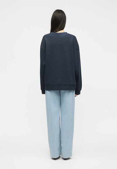Personne aux longs cheveux foncés, de dos, portant un sweat-shirt sombre et oversized ainsi qu’un jean large bleu clair sur un fond uni.