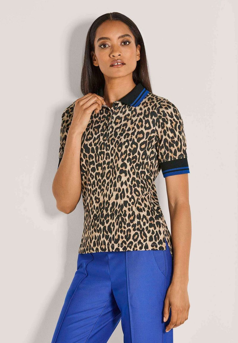 Vrouw met lang donker haar, gekleed in een polo shirt met luipaardprint met zwarte en blauwe accenten, gecombineerd met felblauwe broek, staand tegen een effen muur.