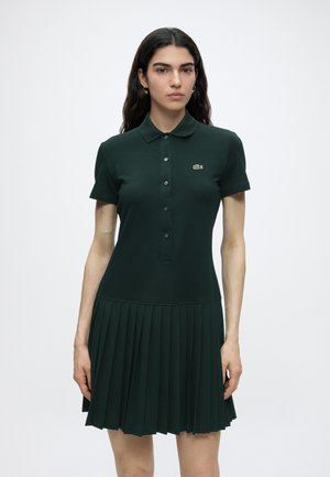 Donna che indossa un abito polo verde scuro a maniche corte con patta con bottoni e gonna plissettata, in piedi su uno sfondo bianco uniforme.