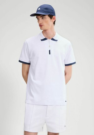 Joven con camisa polo blanca con ribetes en azul marino, pantalones cortos blancos y gorra azul marino, de pie contra un fondo liso claro.