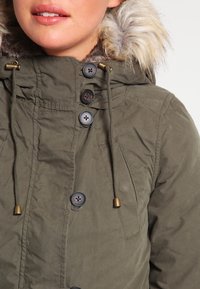 Olivově zelená parka bunda s kapucí lemovanou kožešinou, šňůrkou a několika knoflíky. Má strukturovanou tkaninu a viditelné stehy.