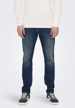 Man draagt verweerde blauwe slim-fit jeans, witte gestructureerde trui en zwarte sneakers met witte strepen, staand tegen een effen achtergrond.