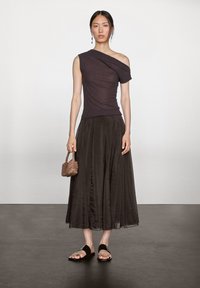 Donna in piedi che indossa un top scuro a spalla unica, una lunga gonna nera, sandali neri e tiene in mano una piccola borsa intrecciata di colore marrone.