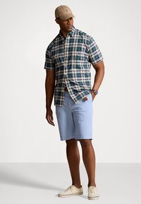 Short-sleeve plaid shirt in green and navy, paired with light blue shorts and beige sneakers. A beige cap complements the outfit.