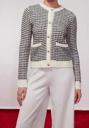 Femme portant un cardigan boutonné rayé noir et blanc avec deux poches avant et un pantalon blanc, debout sur un tapis rouge.