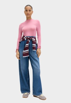 Langærmet lyserød top kombineret med blå denimbukser med vide ben, fremhævet af et maroon og hvidstribet bånd bundet i taljen. Sko i neutrale toner.