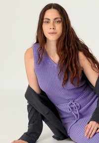 Lila gestricktes ärmelloses Kleid mit strukturiertem Muster und Kordelzug in der Taille, das mit einem schwarzen Blazer über den Schultern getragen wird. Einfaches und tailliertes Design.