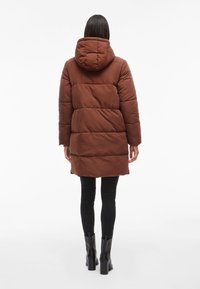 Cappotto puffer marrone con cappuccio, caratterizzato da un design trapuntato e pannelli verticali. Abbinato a leggins neri e stivaletti alla caviglia.