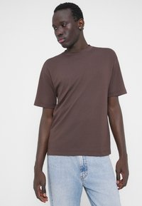 Rjava kratka majica s kratkimi rokavi iz mehke tkanine, z ravnim izrezom in okroglim ovratnikom, v kombinaciji s svetlo modrimi jeansi iz jeansa.