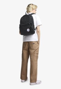 Černý látkový batoh s přední kapsou na zip a logovou nášivkou. Nositel má na sobě bílou košili a béžové nákladní kalhoty, je otočen zády.