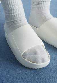 Hvite sandaler med åpen tå og en glatt, glinsende overflate, sammen med hvite ribbestrikkede sokker, plassert på et blått teppe.