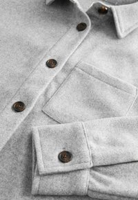 Gray flannel shirt featuring a collar, chest pocket, and brown buttons. Soft texture with straight cuffs and visible stitching details.