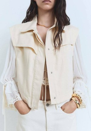 Beige sleeveless jacket made of cotton, featuring large front pockets and snap button closure. Underneath, a white blouse with lace trim.
