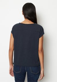 Donna con capelli lunghi e scuri che indossa una t-shirt a maniche corte blu navy e jeans blu scuro, in piedi di fronte a un muro bianco.