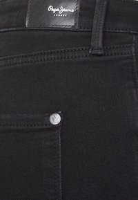 Jeans de mezclilla negra con un parche de cuero, costuras en contraste y un detalle de remache metálico en el bolsillo delantero.