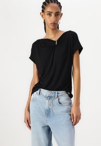 Top corto nero con dettaglio frontale attorcigliato e accento metallico, abbinato a jeans azzurri larghi con cintura a catena.