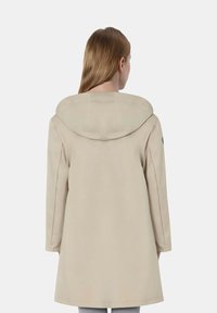 Cappotto beige con un ampio cappuccio, maniche lunghe e taglio dritto. Presenta dettagli di cucitura minima e una texture liscia.