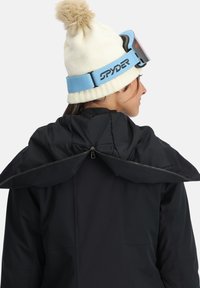 Schwarze Skijacke mit einem Reißverschluss am Rücken und einer weichen, strukturierten Kapuze. Accessoires sind eine cremefarbene Mütze mit Pompon und ein blauer Brillenband.