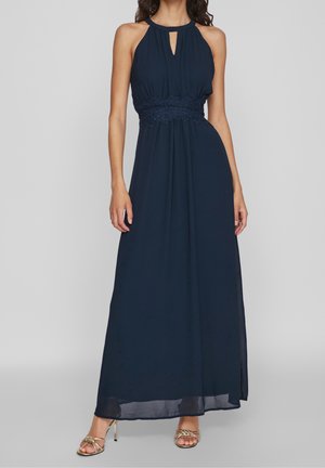 Kvinde iført navyblå maxi-kjole uden ærmer med halterneck, blonder i taljen og guld fletrede stropper på hælene, stående mod grå baggrund.